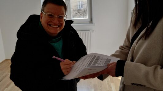Eine Frau mit kurzen Haaren und Brille steht in einer leeren Wohnung und unterschreibt Formulare. Sie übernimmt eine neue Wohnung.