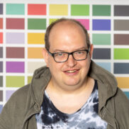 Ein Mann mit Brille schaut freundlich in die Kamera. Er steht vor einer bunt gemusterten WAnd und trägt ein T-Shrit mit einer Jacke darüber