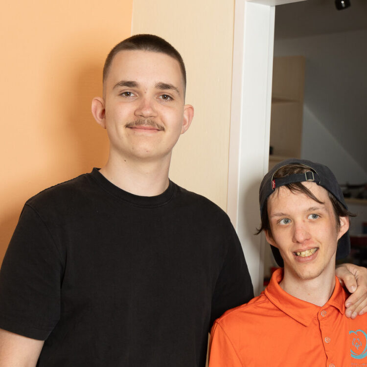 Zwei junge Männer schauen freundlich in die Kamera. Der linke ist ein Zivildiener ganz in schwarz gekleidet, er legt seine Hand auf die Schulter eines Klienten der Lebenshilfe mit orangenem T-Shirt