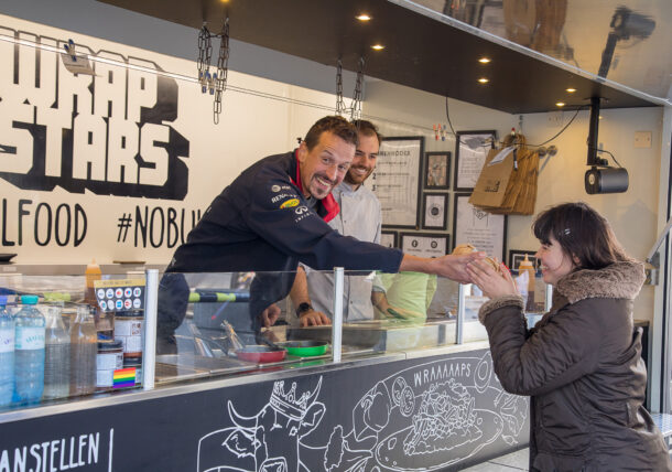 Ein Kunde der Lebenshilfe Wien, ein schlanker Mann mit kurzem Haar, reicht einer Frau aus einem Foodtruck einen Wrap. Der Foodtruck gehört der Firma Wrapstars, einem Kooperationspartner der Lebenshilfe Wien