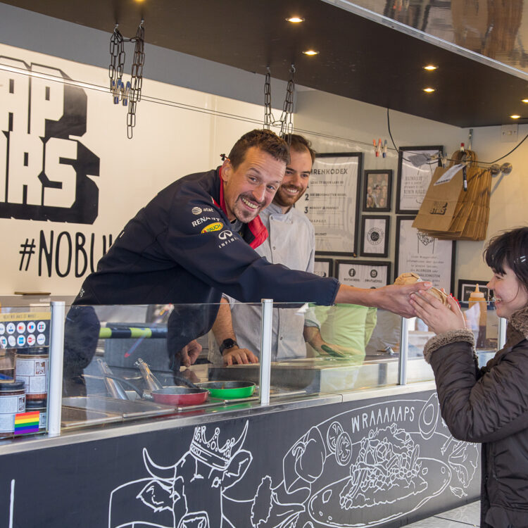 Ein Kunde der Lebenshilfe Wien, ein schlanker Mann mit kurzem Haar, reicht einer Frau aus einem Foodtruck einen Wrap. Der Foodtruck gehört der Firma Wrapstars, einem Kooperationspartner der Lebenshilfe Wien