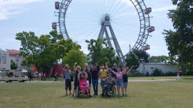 Eine fröhliche Gruppe von Menschen, darunter auch Personen im Rollstuhl, steht auf einer Wiese vor dem Wiener Riesenrad. Alle heben die Arme und lachen. Im Hintergrund sind Bäume, ein kleiner Zug und Spaziergänger zu sehen.