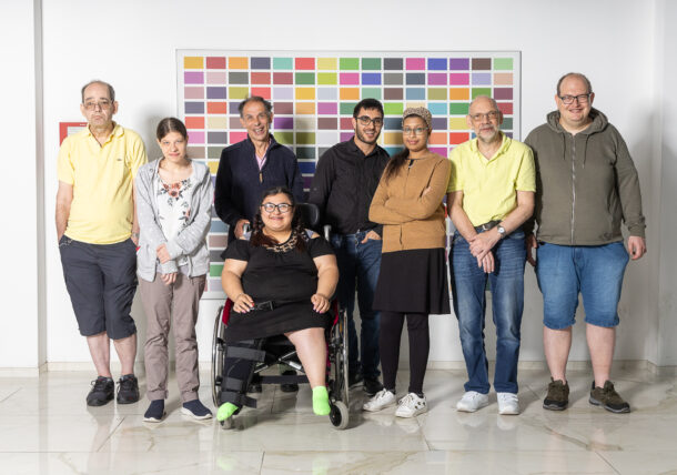 Eine Gruppe von acht Personen steht zusammen vor einer weißen Wand mit einem bunten Rasterbild. In der Mitte sitzt eine Frau im Rollstuhl, die schwarze Kleidung und eine Beinorthese trägt. Sie lächelt und trägt eine Brille. Um sie herum stehen acht weitere Personen, eine diverse Gruppe unterschiedlichen Alters und Geschlechts, einige mit Brillen, manche leger, andere etwas formeller gekleidet. Alle blicken in die Kamera, einige lächeln, andere haben einen neutralen Gesichtsausdruck. Der Boden ist aus hellem Marmor, und die Atmosphäre wirkt freundlich und inklusiv.