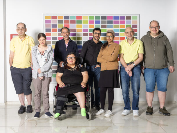 Eine Gruppe von acht Personen steht zusammen vor einer weißen Wand mit einem bunten Rasterbild. In der Mitte sitzt eine Frau im Rollstuhl, die schwarze Kleidung und eine Beinorthese trägt. Sie lächelt und trägt eine Brille. Um sie herum stehen acht weitere Personen, eine diverse Gruppe unterschiedlichen Alters und Geschlechts, einige mit Brillen, manche leger, andere etwas formeller gekleidet. Alle blicken in die Kamera, einige lächeln, andere haben einen neutralen Gesichtsausdruck. Der Boden ist aus hellem Marmor, und die Atmosphäre wirkt freundlich und inklusiv.