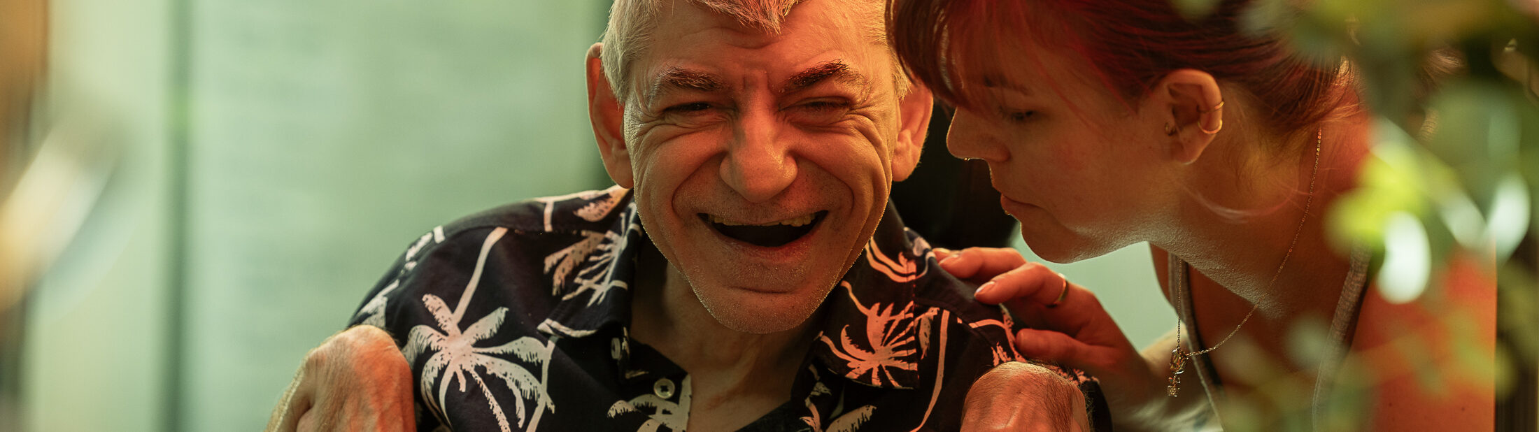 Ein älterer Mann mit kurzen grauen Haaren sitzt in einem Rollstuhl und lacht herzlich. Er trägt ein dunkles Hemd mit einem Palmenmuster. Eine Frau mit roten Haaren beugt sich liebevoll zu ihm, hält ihn sanft und scheint mit ihm zu sprechen. Die Szene ist in warmes Licht getaucht, und im Vordergrund sind leicht verschwommene Pflanzen und Gegenstände zu sehen, die eine gemütliche Atmosphäre schaffen.