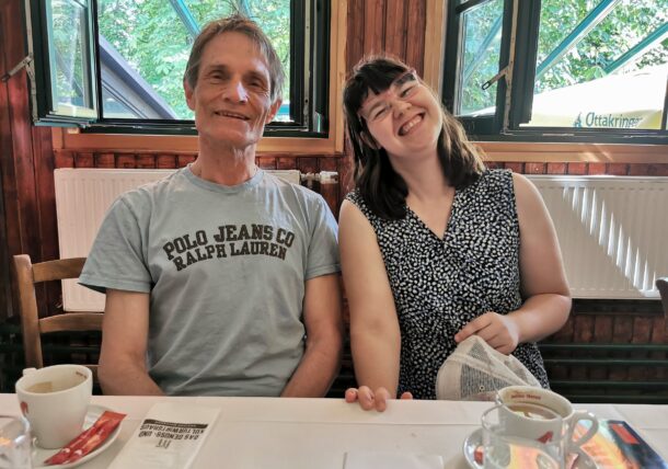 Ein älterer Mann mit brunetten Haaren und grauem Kurzarm-T-Shirt sitzt neben einer jüngeren Frau mit brauen Haaren, Brille und ärmellosen gepunkteten Kleid an einem Tisch mit Kaffeetassen; beide lächeln in die Kamera