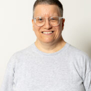 Das Foto zeigt eine Dame mit kurzen Haaren und Brille. Sie lächelt freundlich in die Kamera und trägt ein graues Shirt.