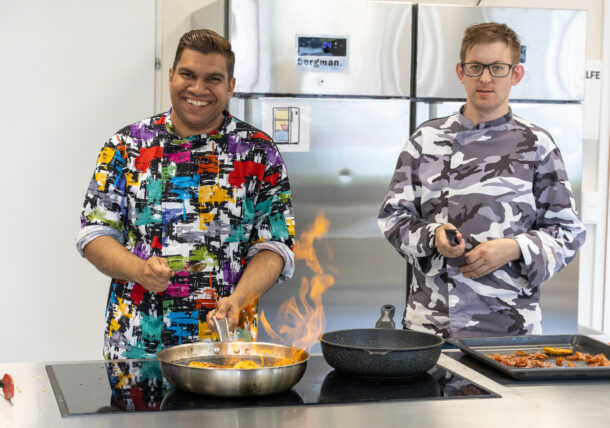 Zwei Männer arbeiten in der Küche, sie tragen bunte Kochjacken und braten Fleisch in einer Pfanne an.