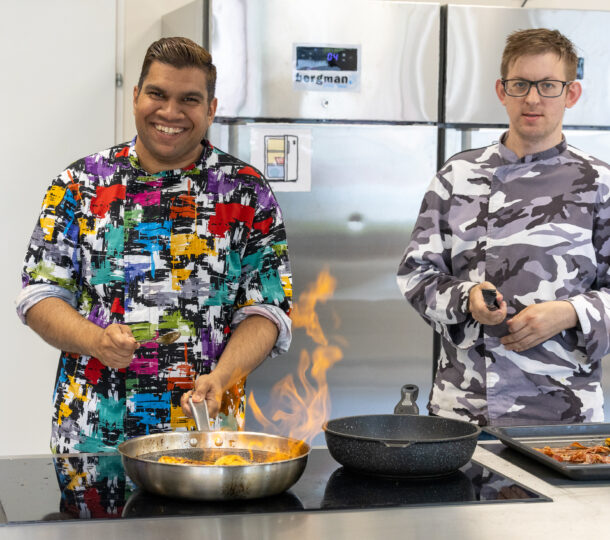 Zwei Männer arbeiten in der Küche, sie tragen bunte Kochjacken und braten Fleisch in einer Pfanne an.