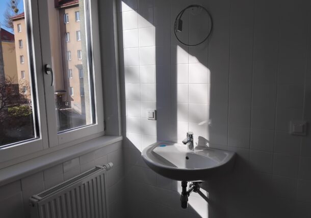 Man sieht einen Teil eines Badezimmers, alles ist neu, weiße Fliesen und weißes Waschbecken. Durch das Fenster scheint die Sonne.