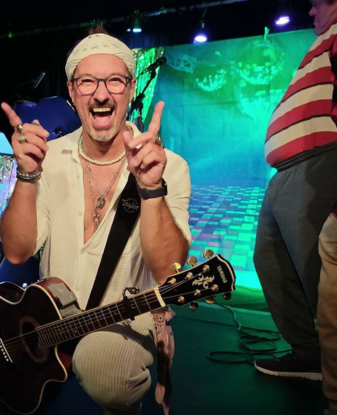 Ein lächelnder Mann mit Bandana und Brille kniet auf der Bühne und hält eine Gitarre in der Hand, umgeben von bunten Bühnenlichtern und anderen Darstellern.