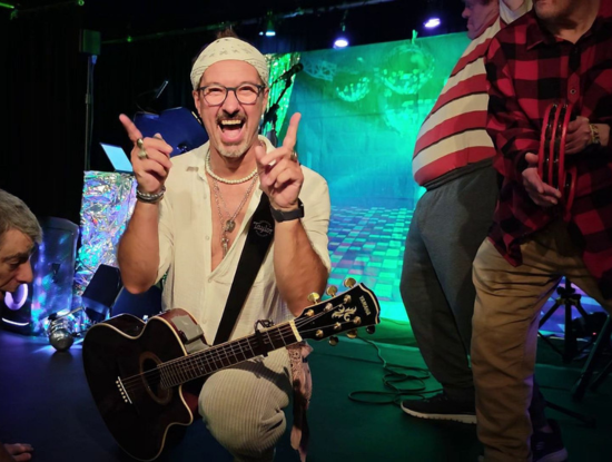 Ein lächelnder Mann mit Bandana und Brille kniet auf der Bühne und hält eine Gitarre in der Hand, umgeben von bunten Bühnenlichtern und anderen Darstellern.