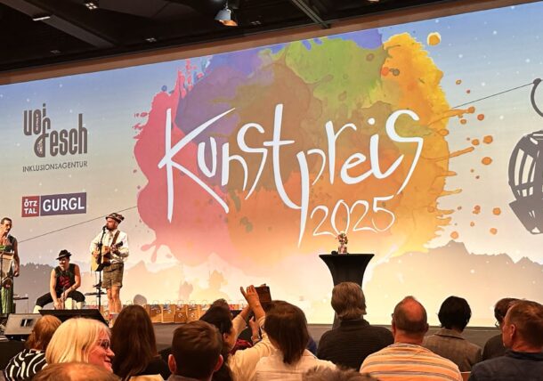 Man sieht eine Veranstaltungsbühne, drei Musiker spielen vor einem Publikum. Im Hintergrund leuchtet das Logo des VOI fesch Kunstpreises 2025 auf einer Leinwand.