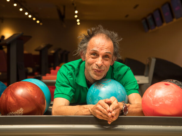 Ein Mann lehnt zwischen bunten Bowling Bällen und schaut freundlich in die Kamera.