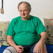 Ein Mann mittleren Alters sitzt in seiner Wohnung auf einer Couch. Er trägt ein grünes T-Shirt