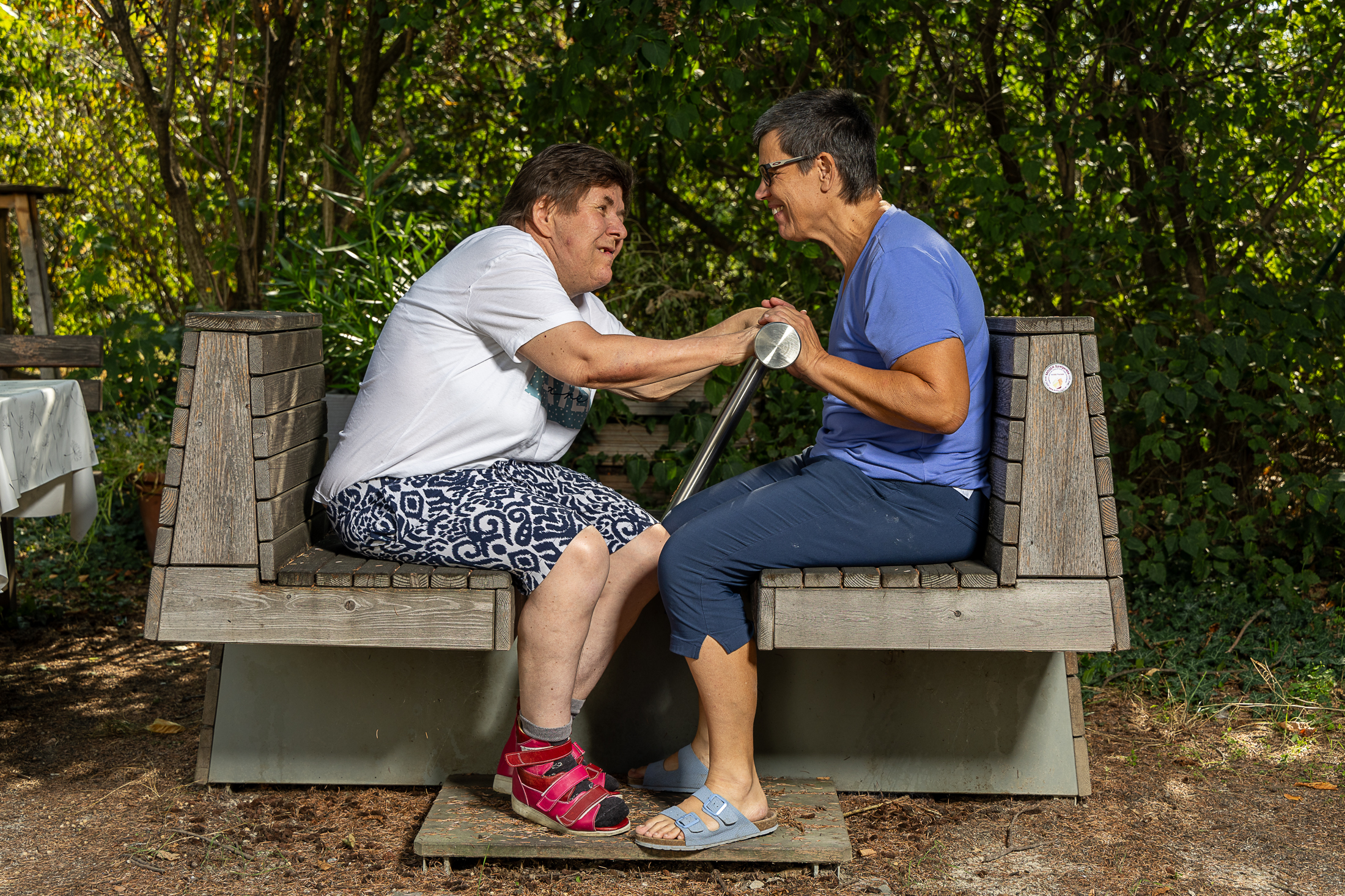 Zwei Frauen in sommerlicher Kleidung sitzen im Garten auf Bänken und machen gemeinsam eine Bewegungsübung. Sie lachen und haben sichtlich Spaß.
