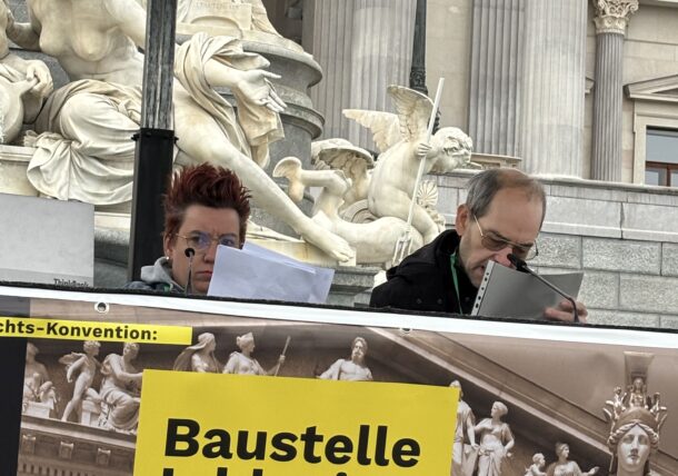Zwei Personen sitzen auf der Bühne vor dem Parlament und lesen aus der UN Behindertenrechts-Konvention vor. Vor ihnen prangt ein Plakat mit der Aufschrift Baustelle Inklusion