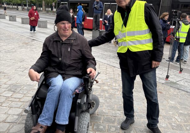 Ein Mann mit einer gelben Warnweste und ein Mann mit Elektro-Rolli stehen vor dem Parlament
