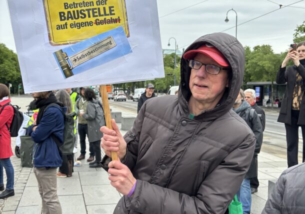 Ein Mann protestiert. Er hat ein Schild in der Hand mit der Aufschrift: Betreten der Baustelle auf eigene Selbstbestimmung