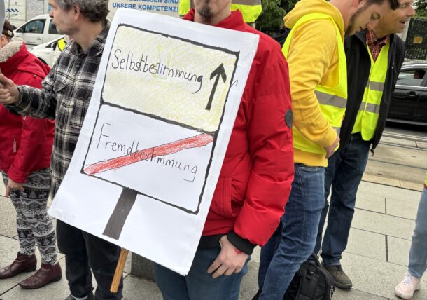 Ein Mann beteiligt sich an der Protestaktion. Er hat ein Schild mit der Aufschrift Selbstbestimmung mit einem Pfeil, der nach oben zeigt. Das Wort Fremdbestimmung ist durchgestrichen