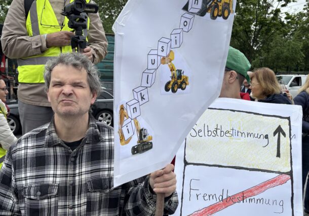 Ein Mann protestiert mit einem Schild, darauf steht Inklusion und man sieht Bagger