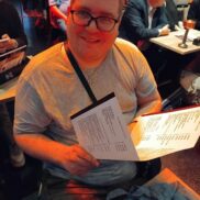 Ein junger Mann mit Brille hält ein Programm in der Hand und lächelt freundlich in die Kamera