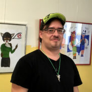 Ein Mann mit Brille und Kappe steht vor seinen Animee Zeichnungen.
