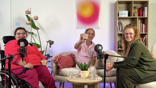 Drei Personen sitzen in einem gemütlichen, gut beleuchteten Raum, in dem Mikrofone für die Aufnahme aufgestellt sind. Eine Person sitzt im Rollstuhl, eine andere hält eine Tasse, und die dritte lächelt. Im Hintergrund stehen Bücherregale und eine Pflanze.