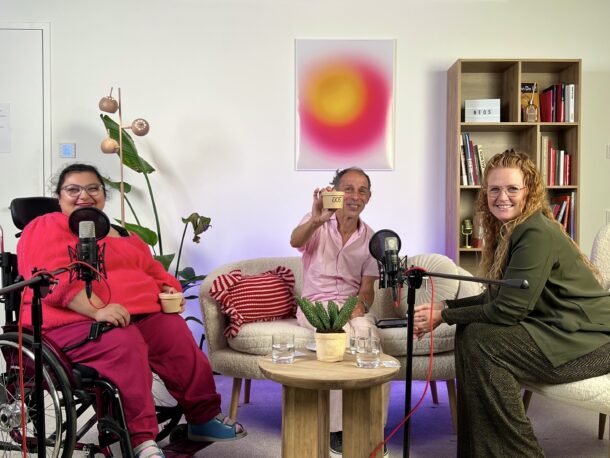 Drei Personen sitzen in einem gemütlichen, gut beleuchteten Raum, in dem Mikrofone für die Aufnahme aufgestellt sind. Eine Person sitzt im Rollstuhl, eine andere hält eine Tasse, und die dritte lächelt. Im Hintergrund stehen Bücherregale und eine Pflanze.