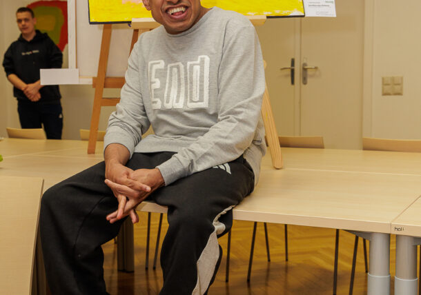 Ein lächelnder Mann in einem grauen Sweatshirt und einer schwarzen Hose sitzt auf der Tischkante vor einem farbenfrohen abstrakten Gemälde auf einer Staffelei bei einer Ausstellung. Eine weitere Person steht im Hintergrund in diesem Innenraum.