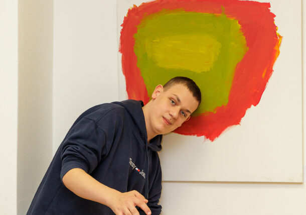 Eine Person in einem marineblauen Kapuzenpulli lehnt mit dem Kopf neben einem farbenfrohen abstrakten Gemälde mit grünen und roten Tönen an einer weißen Wand und gestikuliert spielerisch mit den Händen während einer Ausstellung.