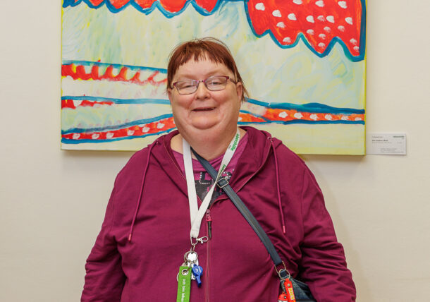 Eine lächelnde Frau mit Brille, kastanienbraunem Kapuzenpulli und einer Umhängetasche steht vor einem farbenfrohen abstrakten Gemälde, das in einer Ausstellung an der Wand hängt.