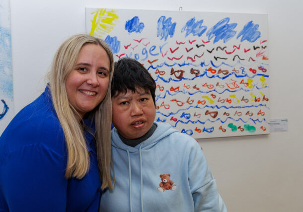 Zwei Personen posieren in der Ausstellung gemeinsam vor einem farbenfrohen, abstrakten Kunstwerk mit Reihen von gemalten Formen und blauen Kritzeleien; eine Person lächelt, während die andere einen neutralen Ausdruck hat.