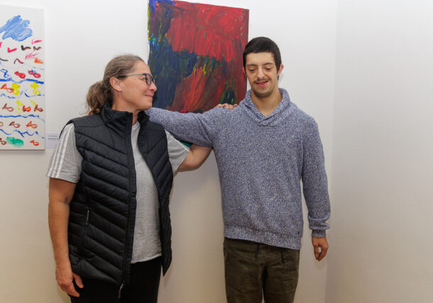 Eine Frau und ein junger Mann stehen lächelnd nebeneinander in einer Kunstgalerie während einer Ausstellung. Die Frau hat ihre Hand auf der Schulter des Mannes. An den weißen Wänden hinter ihnen sind abstrakte Gemälde zu sehen.