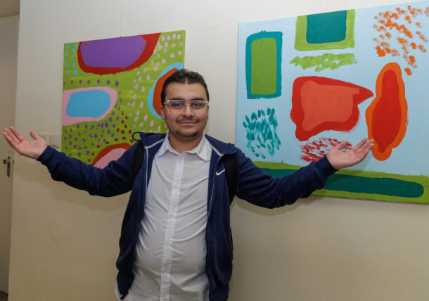 Ein Mann mit Brille und blauer Jacke steht vor zwei farbenfrohen abstrakten Gemälden, lächelt mit offenen Armen und heißt die Besucher der Ausstellung in einer Kunstgalerie willkommen.