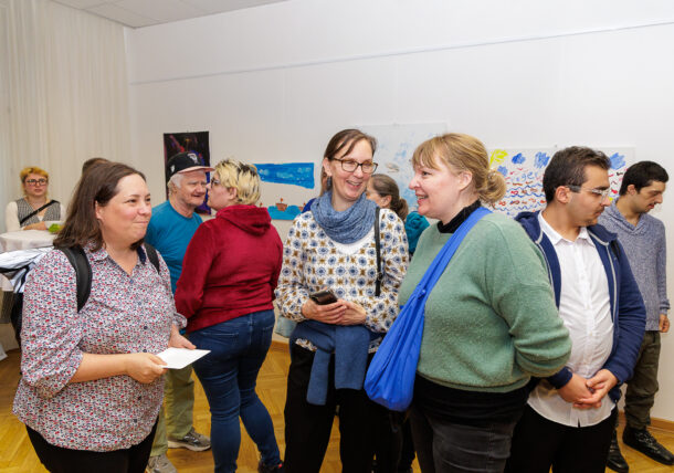 Eine Gruppe von Menschen sitzt während einer Ausstellung in einem hell erleuchteten Raum und lächelt, während an den Wänden Kunst ausgestellt wird. Einige sind in ein Gespräch vertieft, halten Papiere und Telefone in der Hand, was auf eine zwanglose oder gemeinschaftliche Veranstaltung hindeutet.