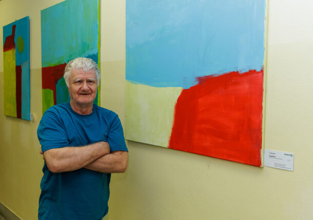 Ein älterer Mann mit grauem Haar und blauem T-Shirt steht mit verschränkten Armen in einem Flur neben abstrakten, farbenfrohen Gemälden, die Teil einer Ausstellung sind.