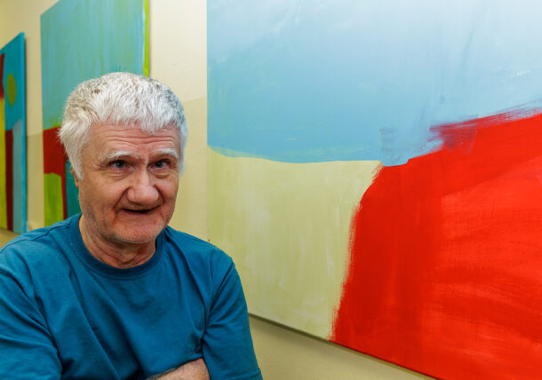 Ein älterer Mann mit grauem Haar und blauem Hemd steht bei einer Ausstellung vor farbenfrohen abstrakten Gemälden, die blaue, rote und cremefarbene Formen an der Wand zeigen.