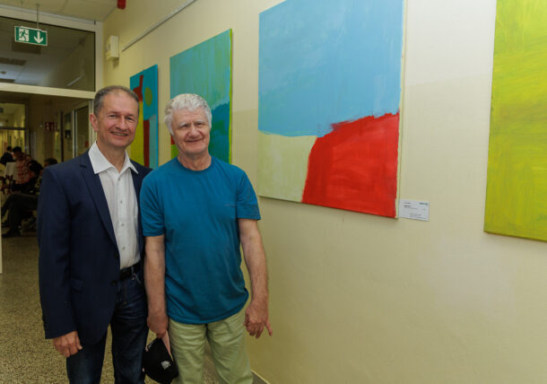 Zwei ältere Männer stehen lächelnd in einem Flur bei einer Ausstellung, hinter ihnen hängen bunte abstrakte Gemälde an der Wand. Der eine trägt eine marineblaue Anzugsjacke und ein weißes Hemd, der andere ein blaues T-Shirt und hält eine dunkle Mütze in der Hand.