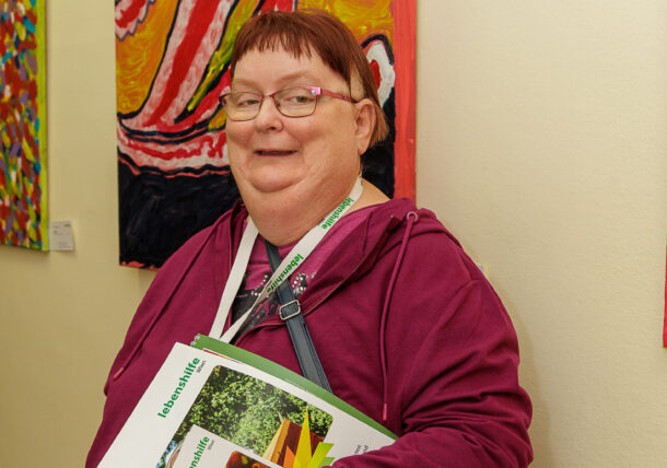 Eine Frau mit Brille und magentafarbenem Kapuzenpulli lächelt, während sie bei einer Ausstellung vor einem farbenfrohen abstrakten Gemälde an einer Galeriewand steht und Broschüren in der Hand hält. Sie trägt eine Umhängetasche und ein Schlüsselband um ihren Hals.