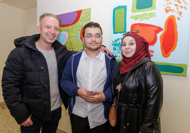 Drei Personen posieren zusammen in einem Innenraum bei einer Ausstellung vor farbenfrohen abstrakten Gemälden. Der Mann links trägt eine schwarze Jacke, der junge Mann in der Mitte trägt eine Brille und ein weißes Hemd, und die Frau rechts trägt einen roten Hidschab und einen schwarzen Mantel.