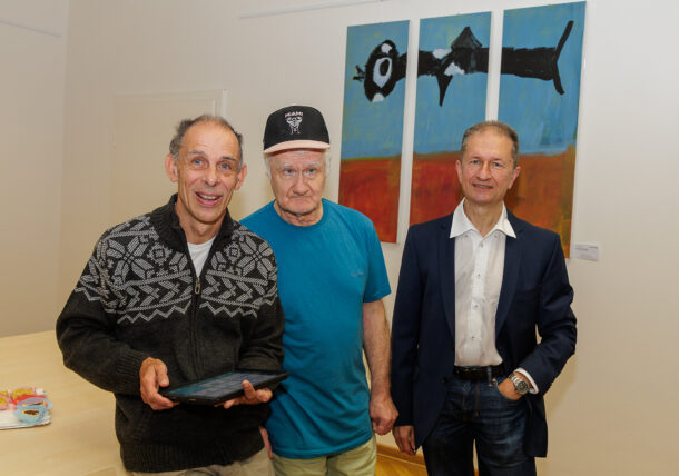 Drei ältere Männer stehen im Innenbereich einer Ausstellung vor farbenfrohen abstrakten Gemälden. Einer hält ein Tablet in der Hand und lächelt, ein anderer trägt ein blaues Hemd und eine weiße Mütze, während der dritte eine Anzugsjacke und ein weißes Hemd trägt, alle posieren für die Kamera.