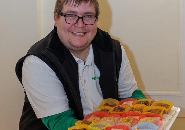 Eine lächelnde Person mit Brille hält ein Tablett mit bunten Cupcakes in Papierverpackungen in der Hand. Sie steht drinnen vor einer weißen Wand und ist bereit, sie bei einer Ausstellung zu präsentieren.