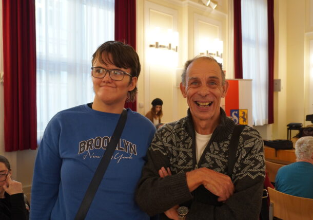 Zwei Personen stehen drinnen in der Ausstellung und lächeln in die Kamera. Einer trägt eine Brille und ein blaues Brooklyn-Sweatshirt, der andere einen gemusterten Pullover. Rote Vorhänge rahmen helle Fenster im Hintergrund ein, in deren Nähe weitere Personen sitzen.