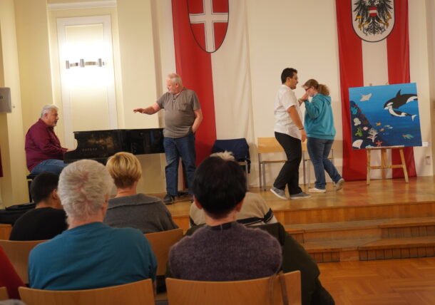 Vier Personen proben auf einer Bühne: Einer spielt Klavier, ein anderer steht und gestikuliert, während zwei eine Szene spielen. Das Publikum schaut von Stühlen aus zu. Hinter ihnen suggerieren große Fahnen und eine Staffelei mit Meerestieren eine Ausstellungsatmosphäre.