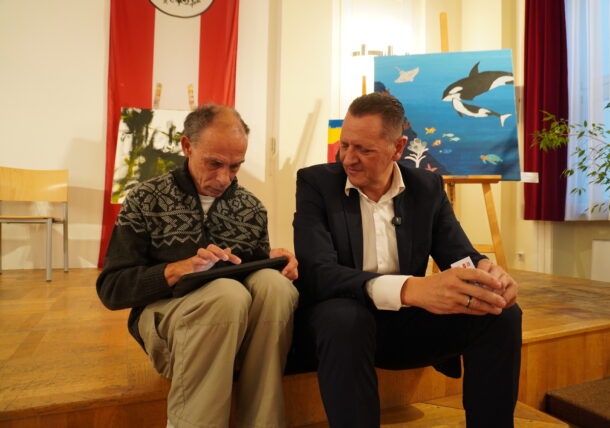In der Ausstellung sitzen zwei Männer auf einer Holzbühne; einer schreibt auf einem Notizblock, während der andere, der einen kleinen Gegenstand in der Hand hält, zusieht. Hinter ihnen stehen Stühle, Tierbilder und eine große rot-weiße Flagge, die an der Wand hängt.