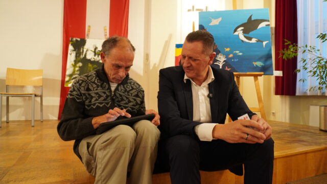 In der Ausstellung sitzen zwei Männer auf einer Holzbühne; einer schreibt auf einem Notizblock, während der andere, der einen kleinen Gegenstand in der Hand hält, zusieht. Hinter ihnen stehen Stühle, Tierbilder und eine große rot-weiße Flagge, die an der Wand hängt.