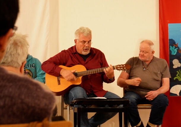 Ein Mann in einem roten Hemd spielt eine Akustikgitarre und singt, während ein anderer Mann in einem grauen Hemd neben ihm sitzt. Sie befinden sich in einem Ausstellungsraum mit einem farbenfrohen Gemälde zum Thema Meer im Hintergrund. Die Zuhörer schauen vom Vordergrund aus zu.