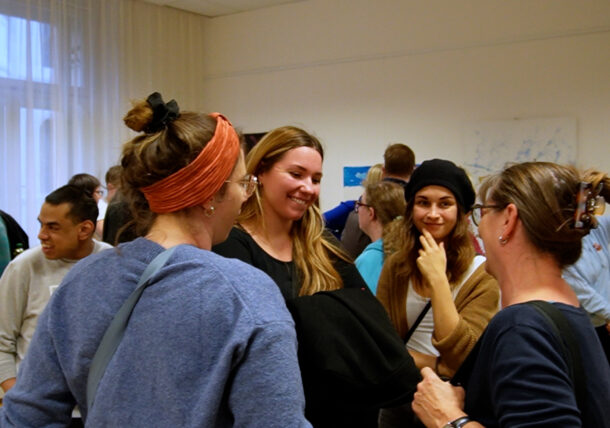 Eine Gruppe von Menschen hat sich im Innenraum der Ausstellung versammelt, unterhält sich und lächelt. Die meisten von ihnen stehen in kleinen Gruppen und unterhalten sich. Die Atmosphäre wirkt zwanglos und freundlich, und an den Wänden sind bunte Kunstwerke zu sehen.