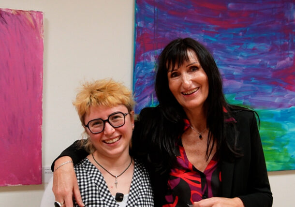 Zwei lächelnde Frauen posieren zusammen vor farbenfrohen abstrakten Gemälden bei einer Ausstellung. Die eine hat kurzes blondes Haar und eine Brille, die andere hat langes dunkles Haar und trägt eine schwarze Jacke. In der Galerie wirken sie glücklich und freundlich.